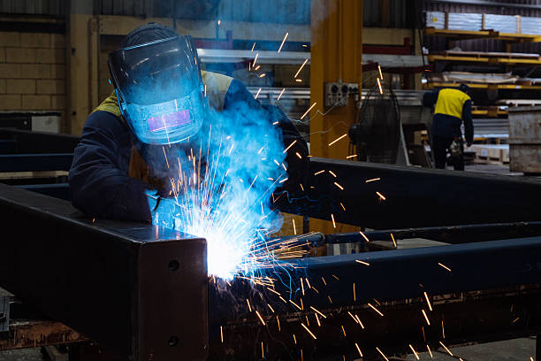 Worker in protective gear welding metal in an industrial factory setting with sparks flying around brightly illuminating the area
